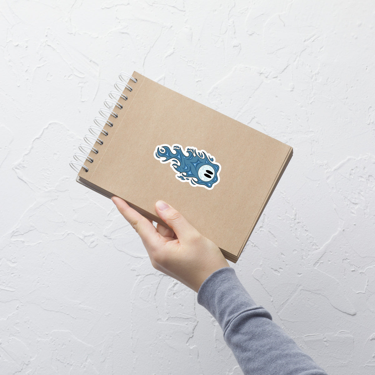 Sticker feu follet bleu 10,2 x 10,2 cm Inari and the Wisps sur un carnet tenu à la main. 